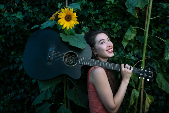 A photo of musician, Miwa Nagato Apthorp smiling radiantly and holding her guitar. Behind her is a blooming sunflower.